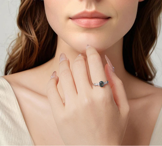 Close-up of a woman's hand wearing a ring with a green gemstone, on a neutral background.


